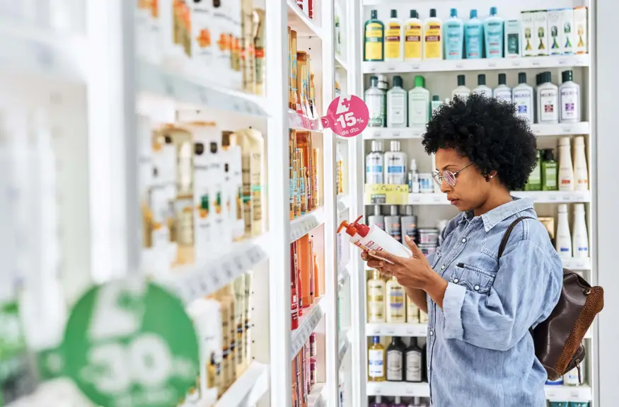 cosmeticos - mulher preta de cabelo crespo vestida de blusa azul clara com mochila marrom nas costas olhando um cosmetico em frente a prateleiras de cosmeticos de uma farmácia